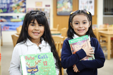 2 elementary school female students in the library holding books and smiling