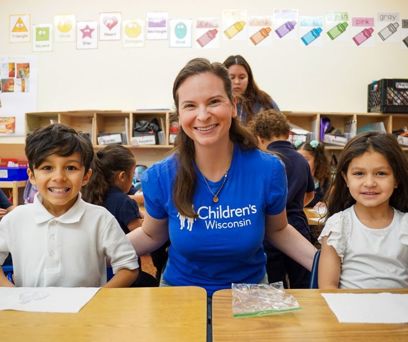 A female volunteer sitting at a desk with 2 elementary aged students smiling